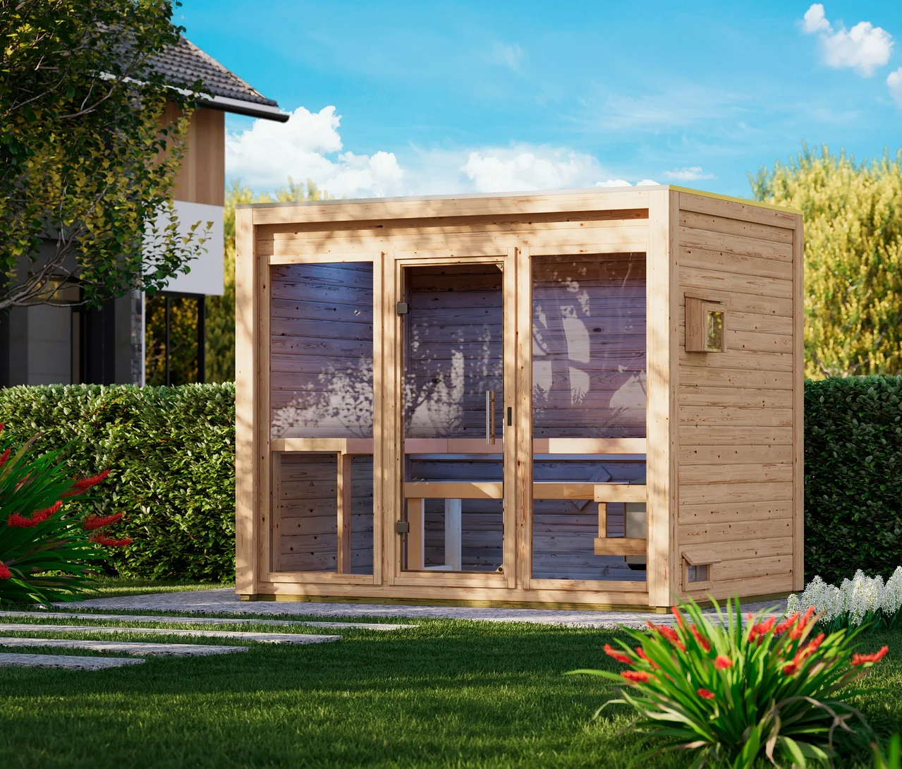 Eine helle Holz-Außensauna mit Glaspaneelen und Glastür steht auf einem gepflasterten Bereich in einem grünen Garten. Im Inneren sind Holzbänke sichtbar. Grüner Rasen, Hecken und rote sowie weiße Blumen umgeben sie, mit einem Haus und Bäumen im Hintergrund unter blauem Himmel.