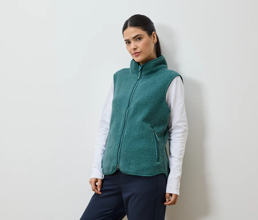 Frau mit Pferdeschwanz, die eine grüne Fleeceweste über einem weißen Langarmshirt und einer marineblauen Hose trägt.