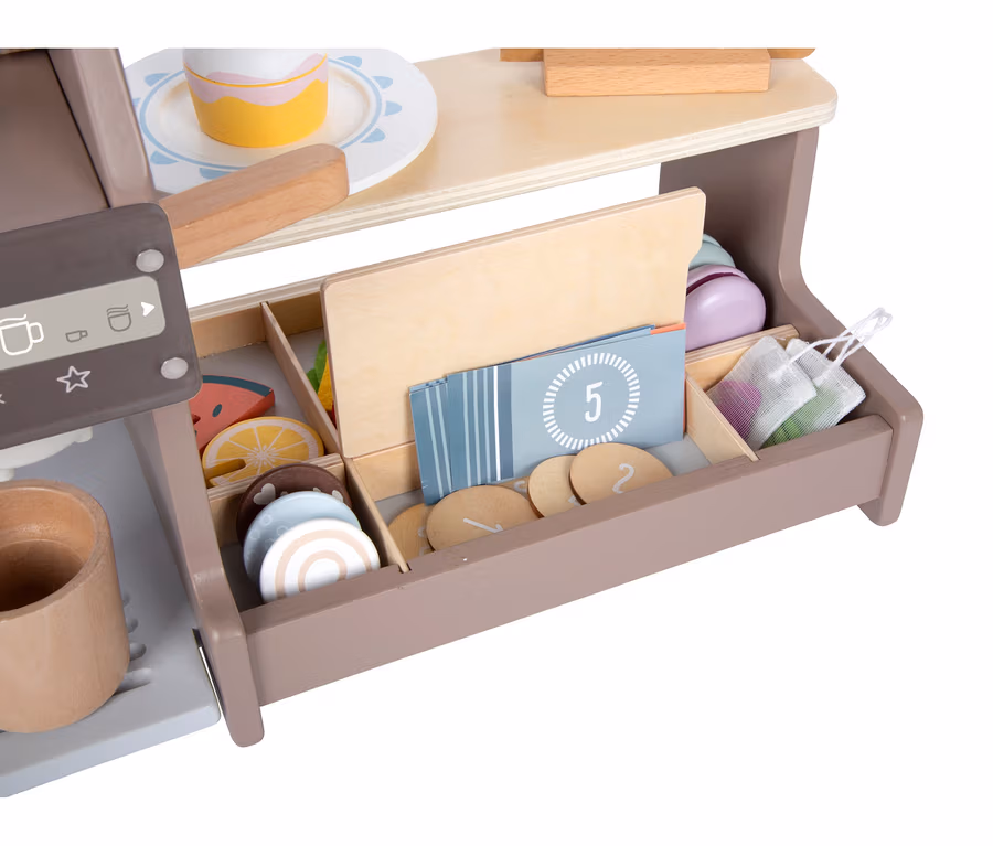 Nahaufnahme einer hölzernen Spielzeug-Kaffeemaschine mit Tasse, einem Kuchen auf einem Teller und einer Schublade voller Spielzeug-Obstscheiben, Kekse, nummerierter Karten und Netzbeutel.