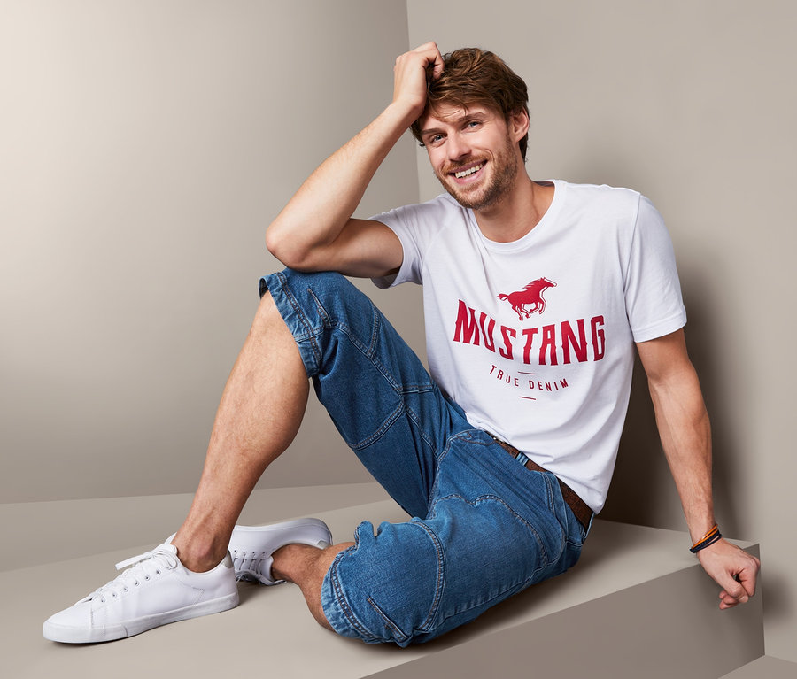 Mann sitzt und trägt Jeans-Bermuda-Shorts »Mustang« und ein T-Shirt.