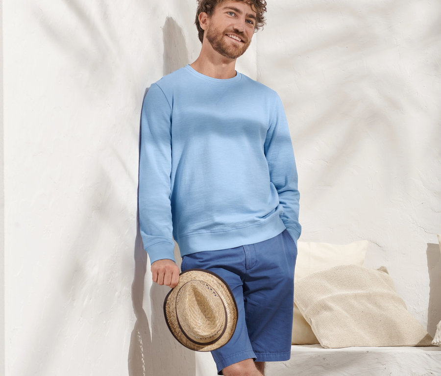 Mann lehnt an Wand, hält Hut und trägt hellblaues Sweatshirt.