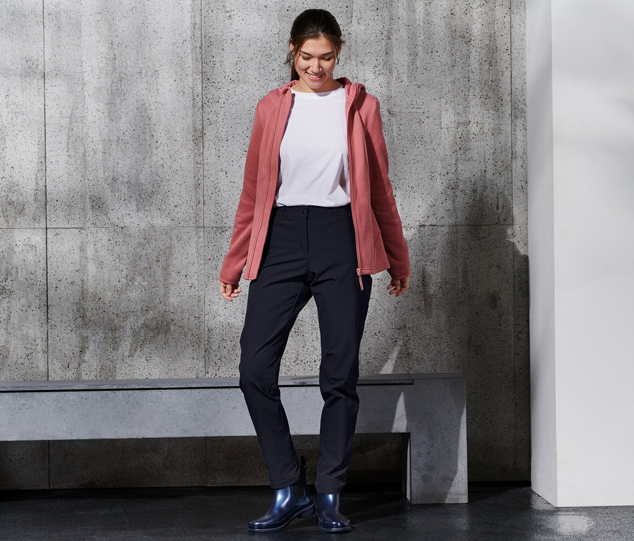 Lächelnde Frau trägt eine Softshell-Hose, ein weißes T-Shirt und eine rosafarbene Kapuzenjacke.