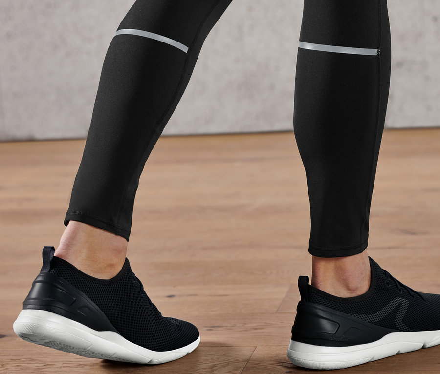 Beine einer Person in schwarzen Performance-Lauftights und schwarzen Schuhen mit weißen Sohlen auf einem Holzboden.