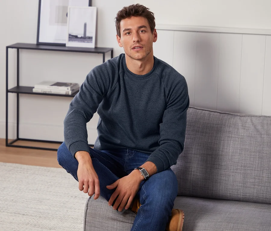 Ein Mann sitzt auf einem grauen Sofa und trägt einen blauen Strickfleecepullover und Jeans.