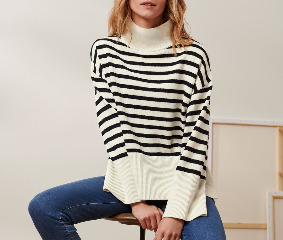 Frau sitzt auf einem Hocker und trägt einen Strickpullover mit Wolle, Jeans und Stiefel.