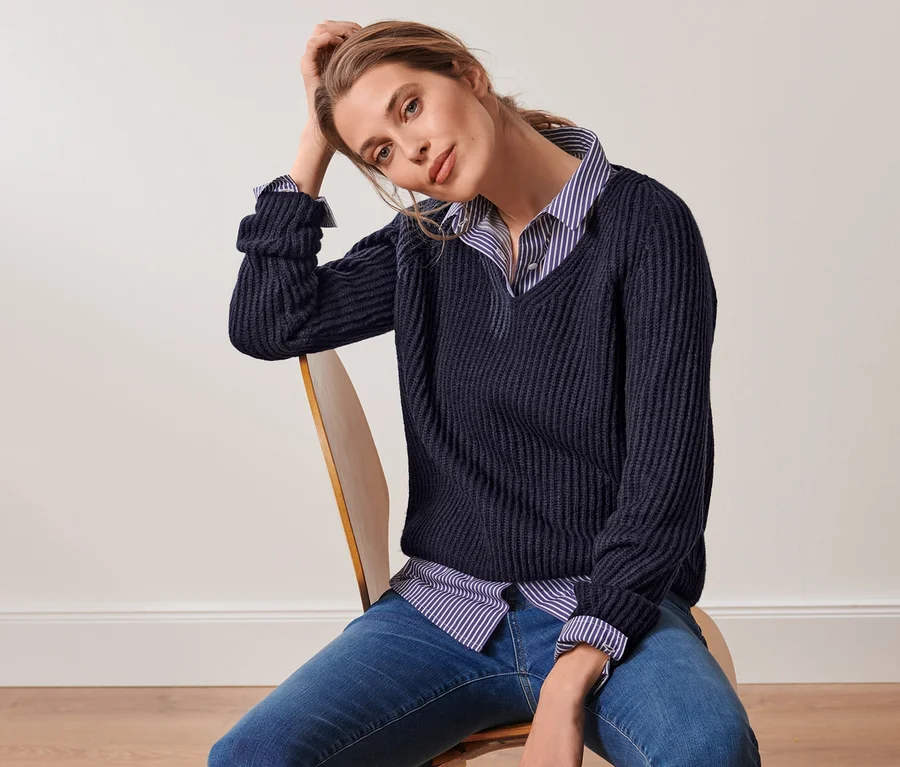 Eine Frau sitzt auf einem Stuhl und trägt eine gestreifte Hemdbluse und einen dunkelblauen Grobstrick-Pullover mit Wolle.
