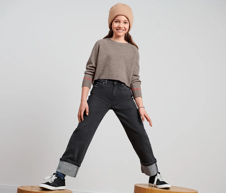 Lächelndes junges Mädchen steht auf Holzhockern und trägt eine Bootcut-Jeans, einen braunen Pullover und eine braune Mütze.