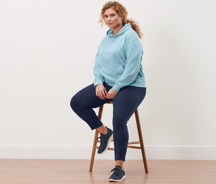 Eine Frau sitzt auf einem Hocker und trägt einen hellblauen Hoodie, eine dunkelblaue Stretchhose und Slip-on-Sneaker.