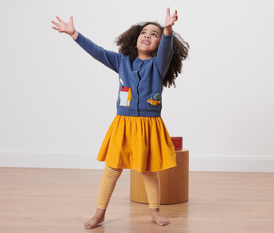 Mädchen mit lockigem Haar steht mit erhobenen Händen. Sie trägt eine blaue Strickjacke mit Applikationen, einen gelben Rock und 3 Kinder-Thermoleggings, Streifen.