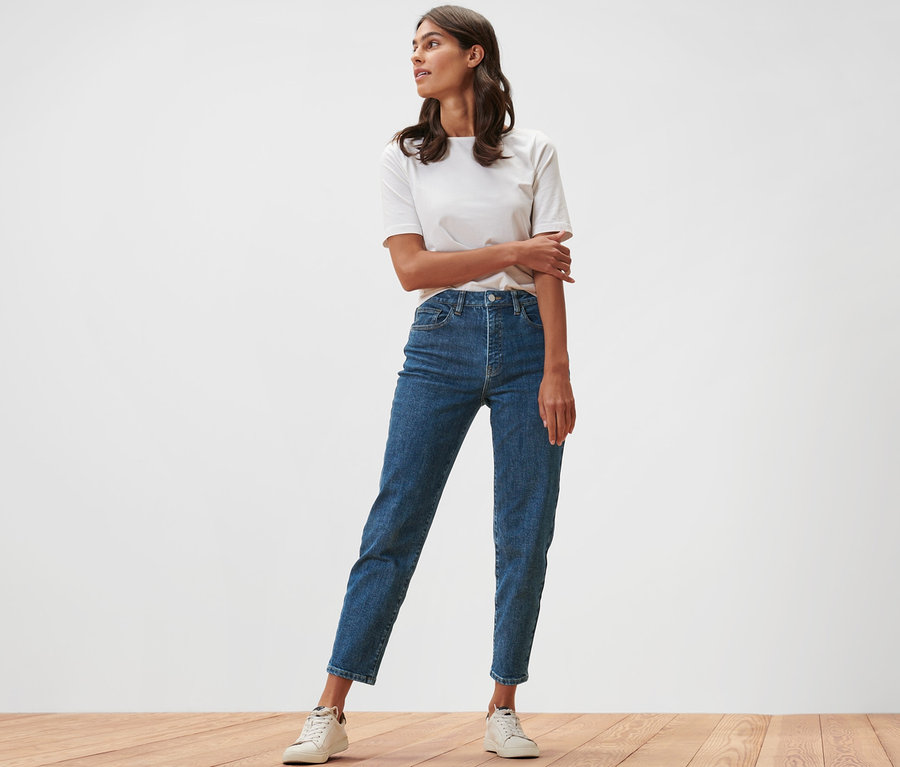 Eine Frau steht auf einem Holzboden und trägt Mom Jeans – Fit »Nyla« und ein weißes T-Shirt.