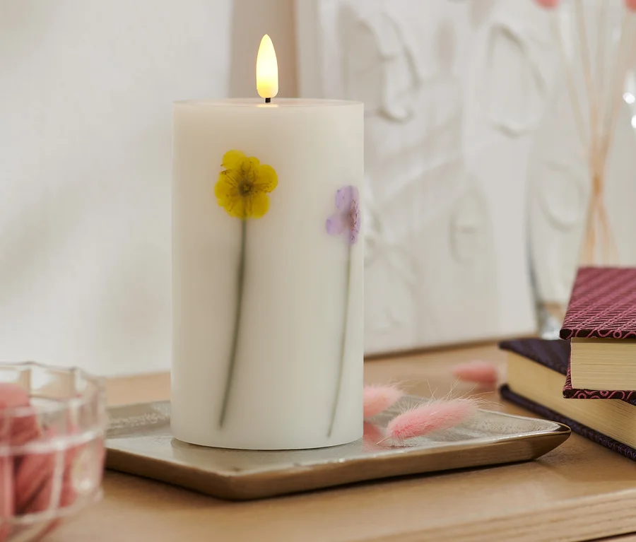 Eine LED-Echtwachskerze mit Trockenblumen steht auf einem Tablett. Im Hintergrund Bücher und eine Vase.