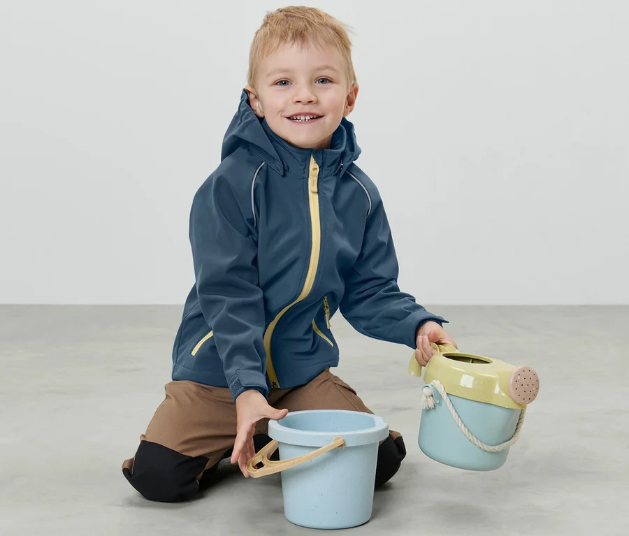 Ein Junge kniet in einer Kleinkind-Softshelljacke auf dem Boden und hält Spielzeug.