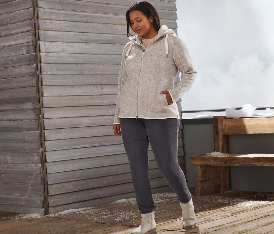 Eine Frau steht draußen und trägt eine beige Teddy-Strickfleecejacke, Slimfit-Jeans und Veloursleder-Boots.