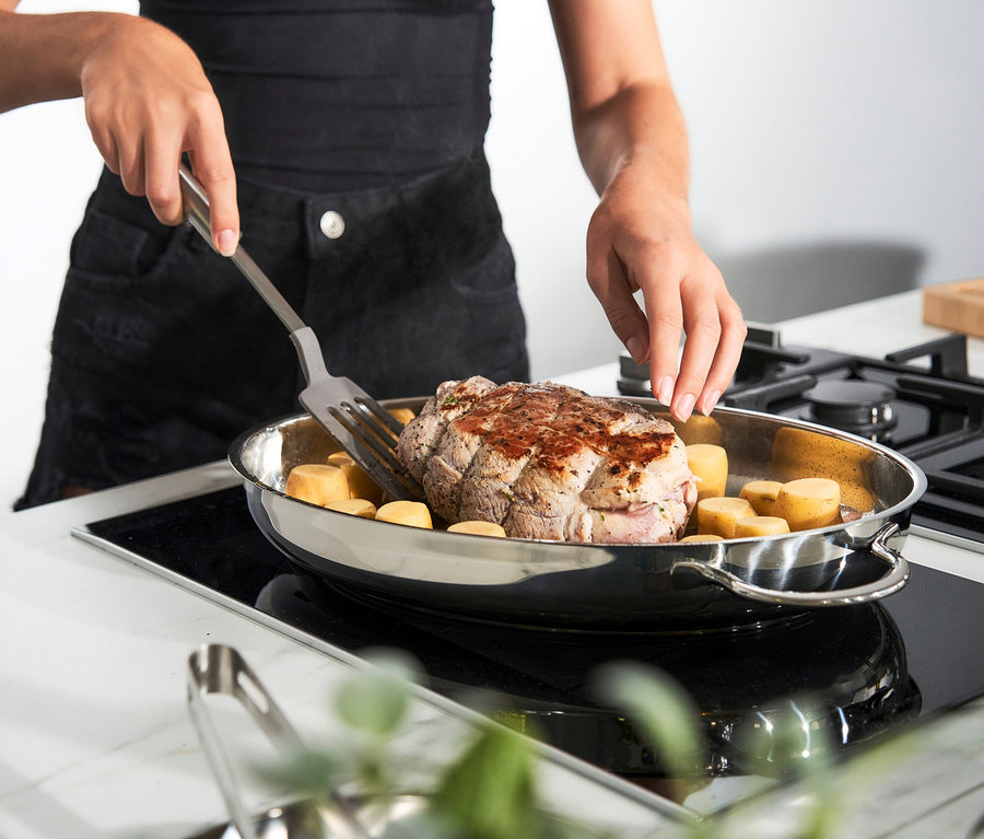 Frau wendet Fleisch mit Kartoffeln in einer ovalen Edelstahlpfanne auf einem Kochfeld.
