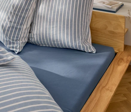 Blaues Spannbetttuch auf Holzbett mit gestreiften Kissen.