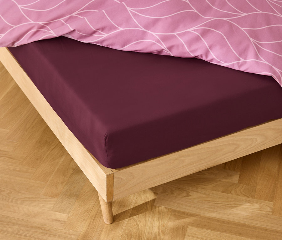 Nahaufnahme eines Bettes mit einem bordeauxroten Spannbetttuch und rosafarbener Bettwäsche mit Muster.