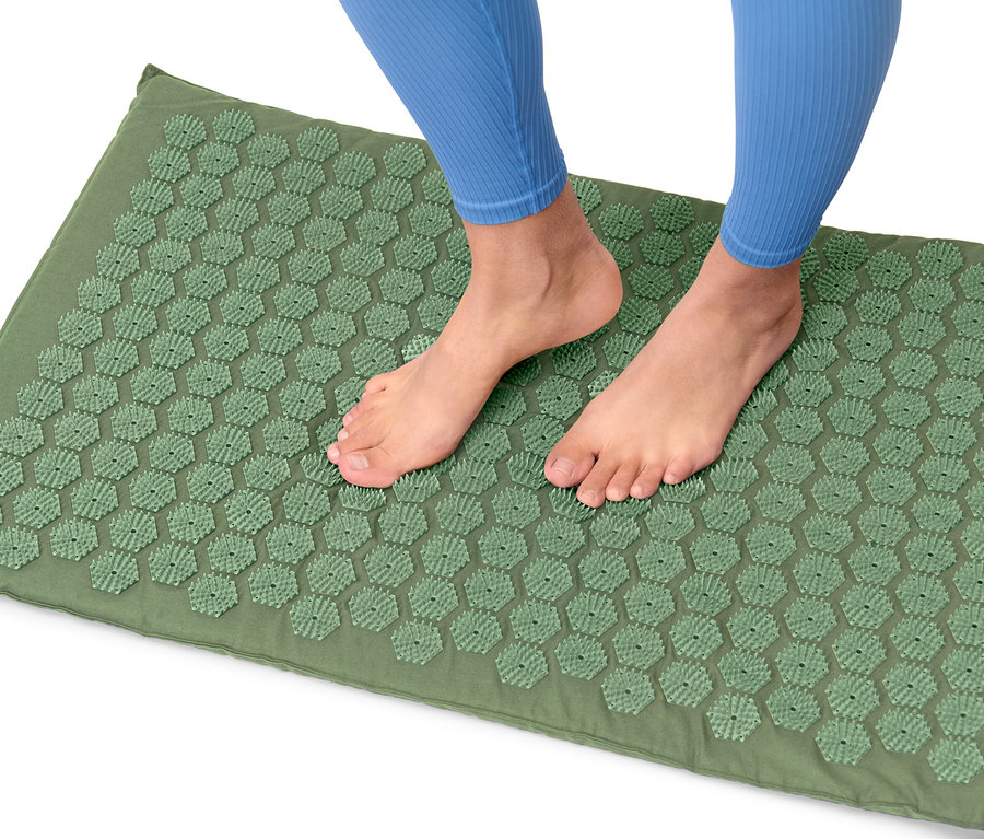 Füße einer Person in blauen Leggings stehen auf einer grünen Akupressurmatte.