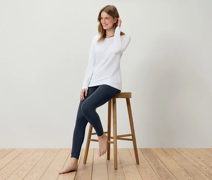 Junge Frau sitzt auf einem Holzhocker, trägt ein weißes Langarmshirt und dunkelblaue Leggings.