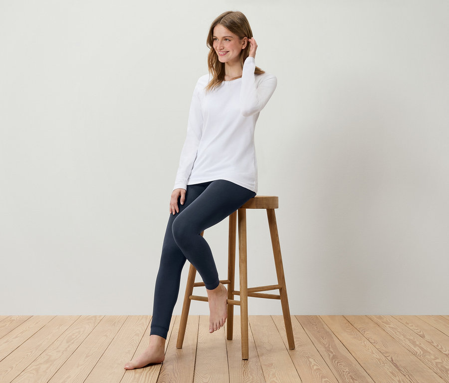 Junge Frau sitzt auf einem Holzhocker, trägt ein weißes Langarmshirt und dunkelblaue Leggings.