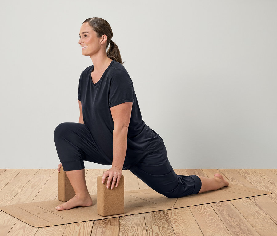 Frau in schwarzer Sportbekleidung macht einen Ausfallschritt auf einer braunen Yogamatte mit Blöcken.