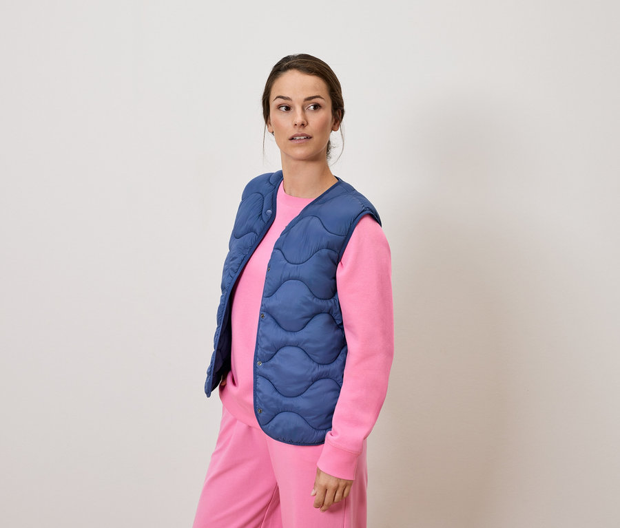 Frau in rosa Anzug und blauer Steppweste steht und schaut in die Ferne.