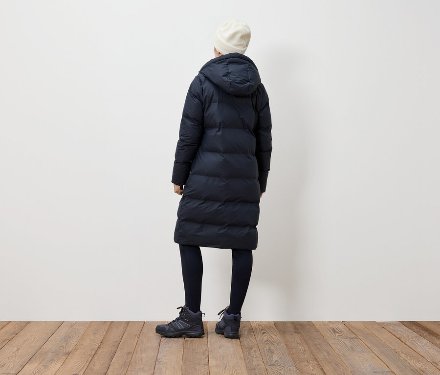 Frau mit dem Rücken zur Kamera trägt einen schwarzen Daunenmantel, schwarze Leggings, schwarze Stiefel und eine weiße Mütze.