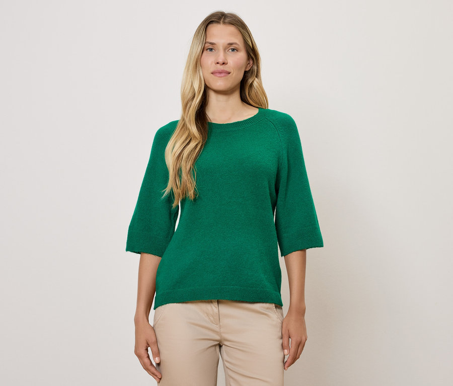 Blonde Frau trägt einen grünen Pullover mit Dreiviertelärmeln und eine beige Hose.