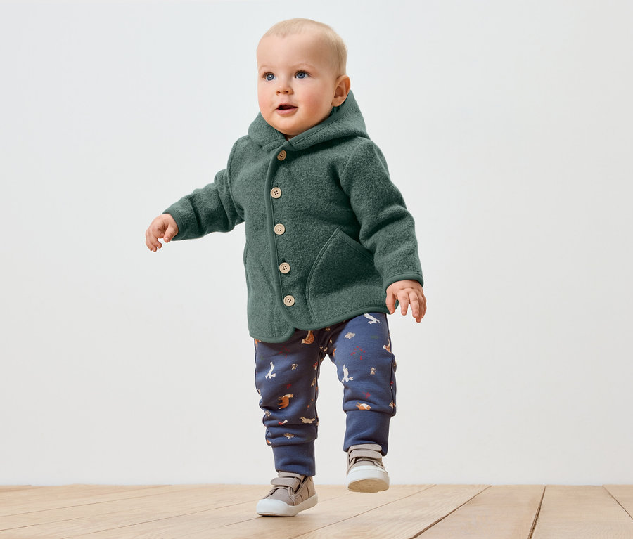 Kleinkind in grüner Wolljacke mit Kapuze und gemusterter Hose macht erste Schritte auf Holzboden.