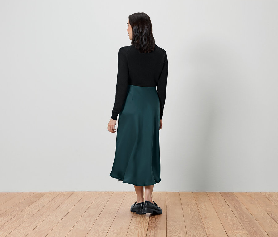 Frau steht mit dem Rücken zur Kamera und trägt ein schwarzes Oberteil und einen grünen Rock.