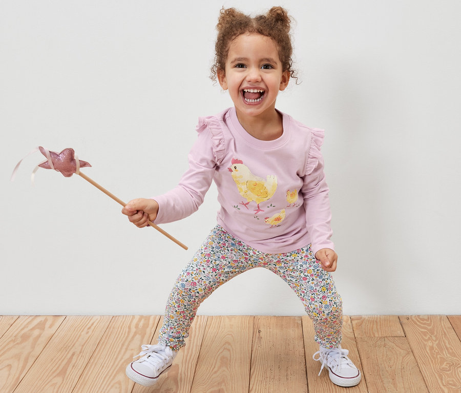Lachendes Mädchen hält einen Zauberstab und trägt eine rosa 2 Kinder-Leggings, rosa und ein T-Shirt mit Hühnerprint.