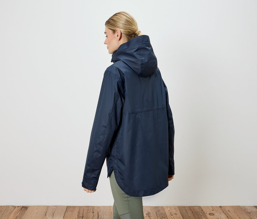 Rückansicht einer Frau mit blonden Haaren, die eine dunkelblaue Regenjacke mit Kapuze und grüne Hosen trägt.