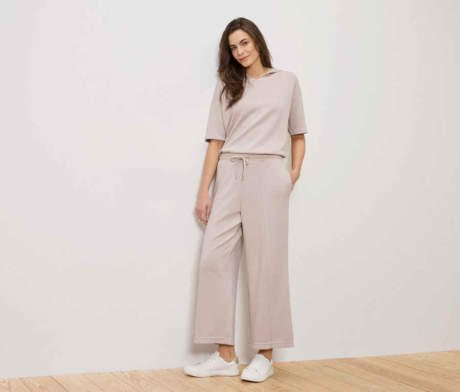 Eine Frau steht vor einer weißen Wand und trägt ein beiges Outfit und weiße Turnschuhe.