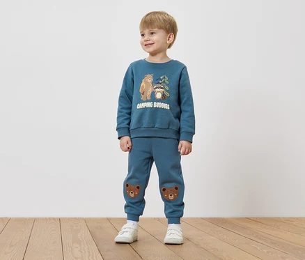 Ein Junge mit blonden Haaren trägt einen blauen Pullover mit Bären- und Waschbär-Motiv und blaue Hosen mit Bärenflicken auf den Knien.