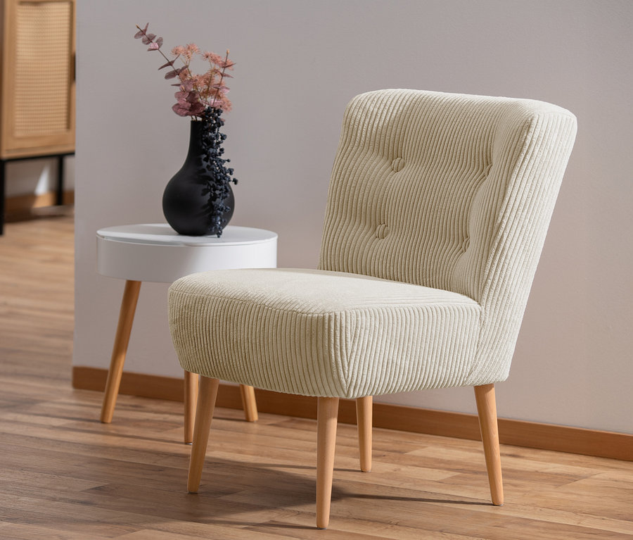 Ein cremefarbener Cocktail-Sessel »Fiona« aus Cordstoff steht auf einem Holzboden. Neben dem Sessel steht ein weißer Beistelltisch mit einer schwarzen Vase.
