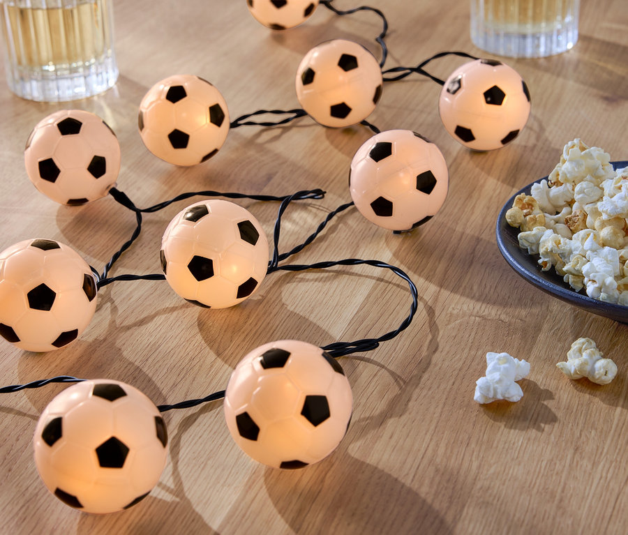 Eine Lichterkette aus fußballförmigen Lichtern liegt auf einem Holztisch neben einer Schale Popcorn und zwei Gläsern mit Getränken.