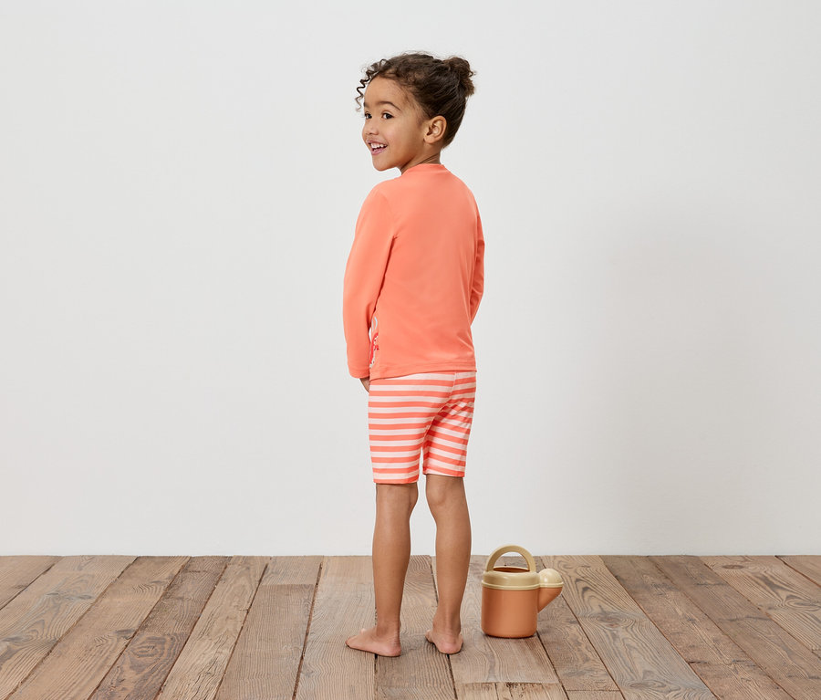Mädchen mit orangem Shirt und gestreiften Shorts, neben einer Gießkanne auf einem Holzboden.
