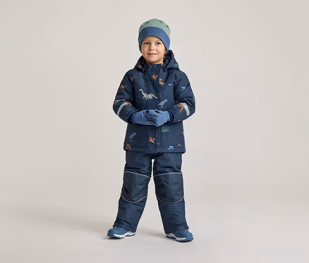 Ein Junge in marineblauer Winterkleidung mit Dinosaurier-Muster, Mütze und Handschuhen steht vor einem hellen Hintergrund.
