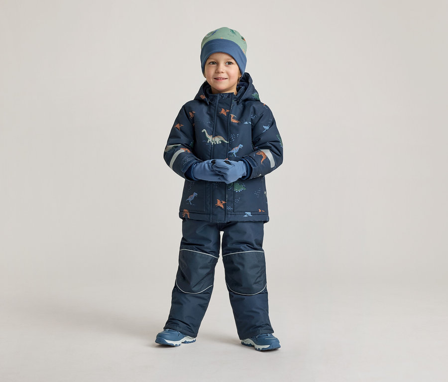 Ein Junge in marineblauer Winterkleidung mit Dinosaurier-Muster, Mütze und Handschuhen steht vor einem hellen Hintergrund.
