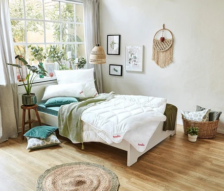 Ein Bett mit einer weißen Paradies Sommerdecke »BestCool« in Normalgröße und Übergröße, Kissen und einer grünen Decke steht vor einem Fenster.