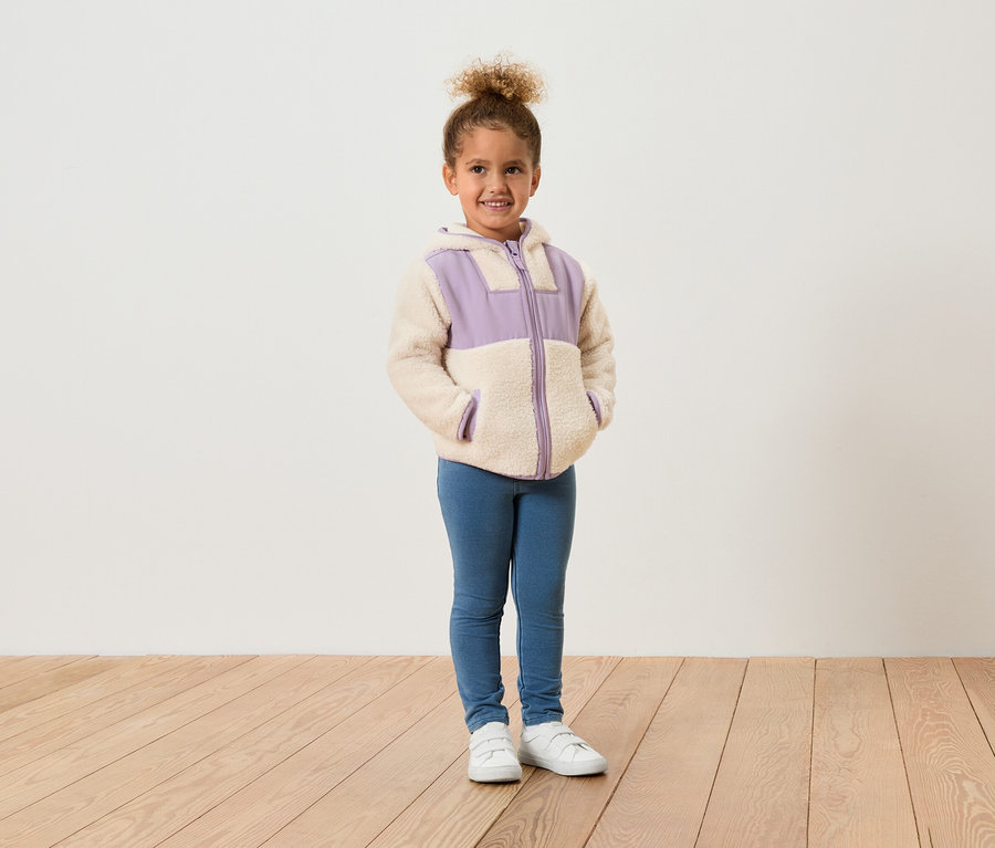 Ein junges Mädchen steht vor einer weißen Wand und trägt eine Fleecejacke mit lila Akzenten, blaue Jeans und weiße Schuhe.