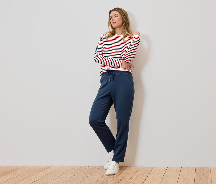 Eine Frau steht vor einer Wand und trägt ein rot-weiß gestreiftes Langarmshirt und eine blaue Jogginghose.