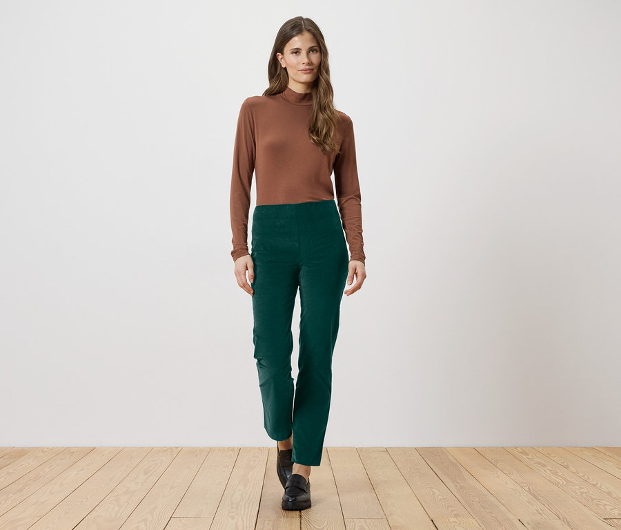 Eine Frau mit langen braunen Haaren trägt ein braunes Langarmshirt und eine grüne Hose. Sie steht auf einem Holzboden.