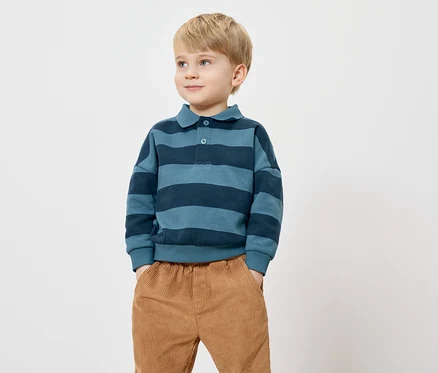Ein Junge mit blonden Haaren steht mit den Händen in den Taschen vor einem weißen Hintergrund. Er trägt ein blau gestreiftes Poloshirt und Cordhosen.