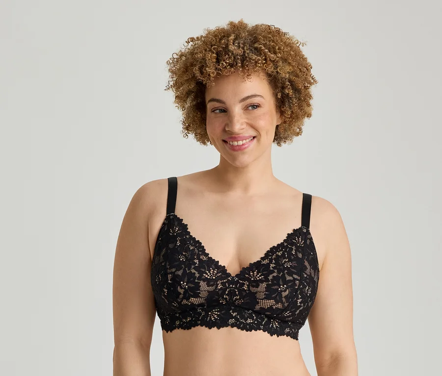 Eine lächelnde Frau mit hellbraunen Locken trägt einen schwarzen Spitzen-Bralette vor einem hellgrauen Hintergrund.