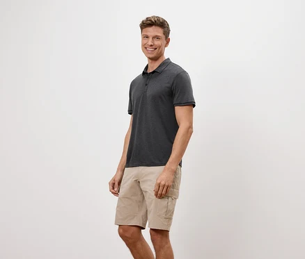 Ein Mann steht und lächelt, er trägt ein anthrazitfarbenes Poloshirt und beigefarbene Shorts.