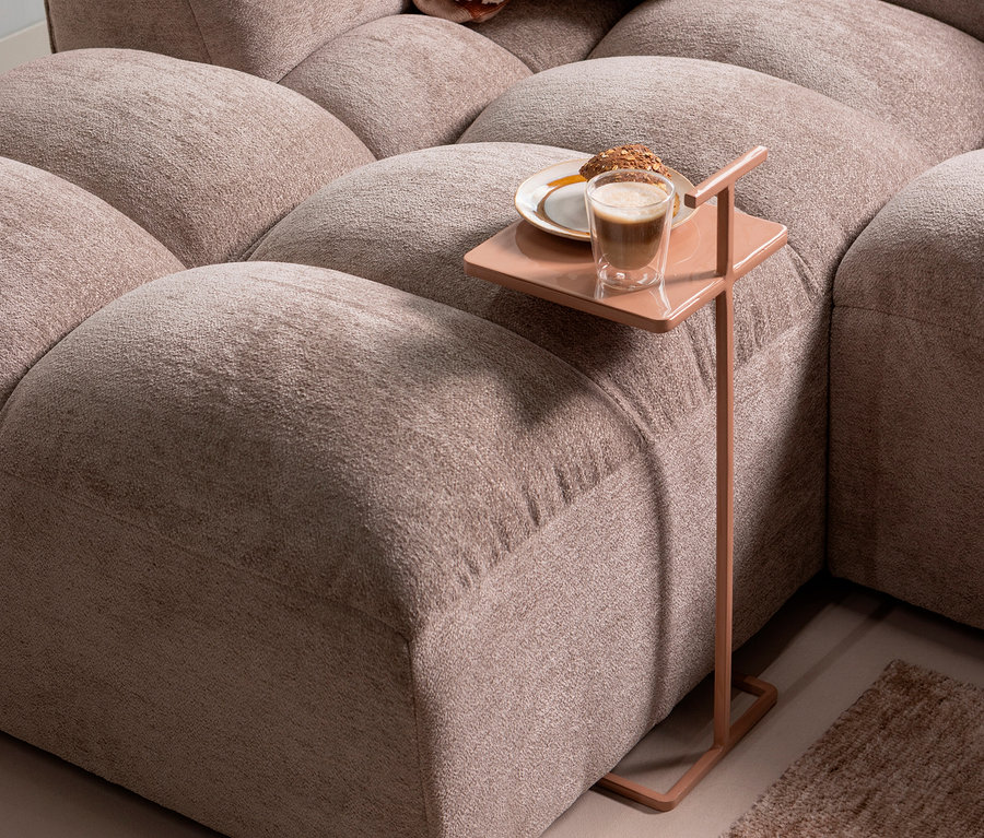 Auf einem kleinen, rosafarbenen Beistelltisch neben einem beige Modulsofa stehen ein Glas Kaffee und ein Teller mit einem Gebäckstück.