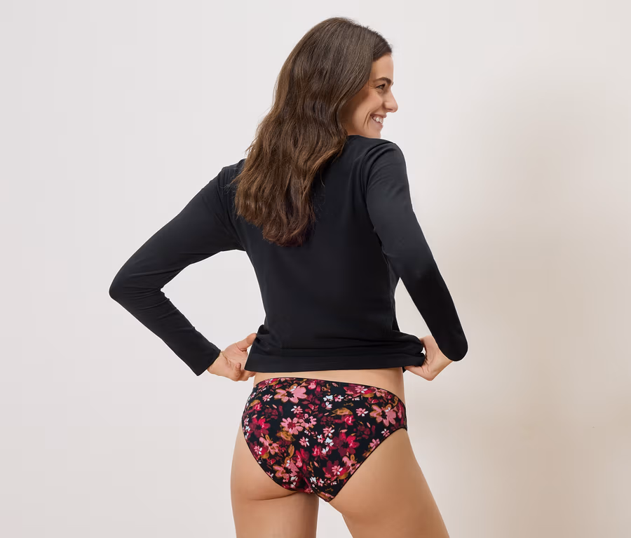 Frau mit dunklen, langen Haaren, die ein schwarzes Langarmshirt und eine Unterhose mit Blumenmuster trägt und lächelt.