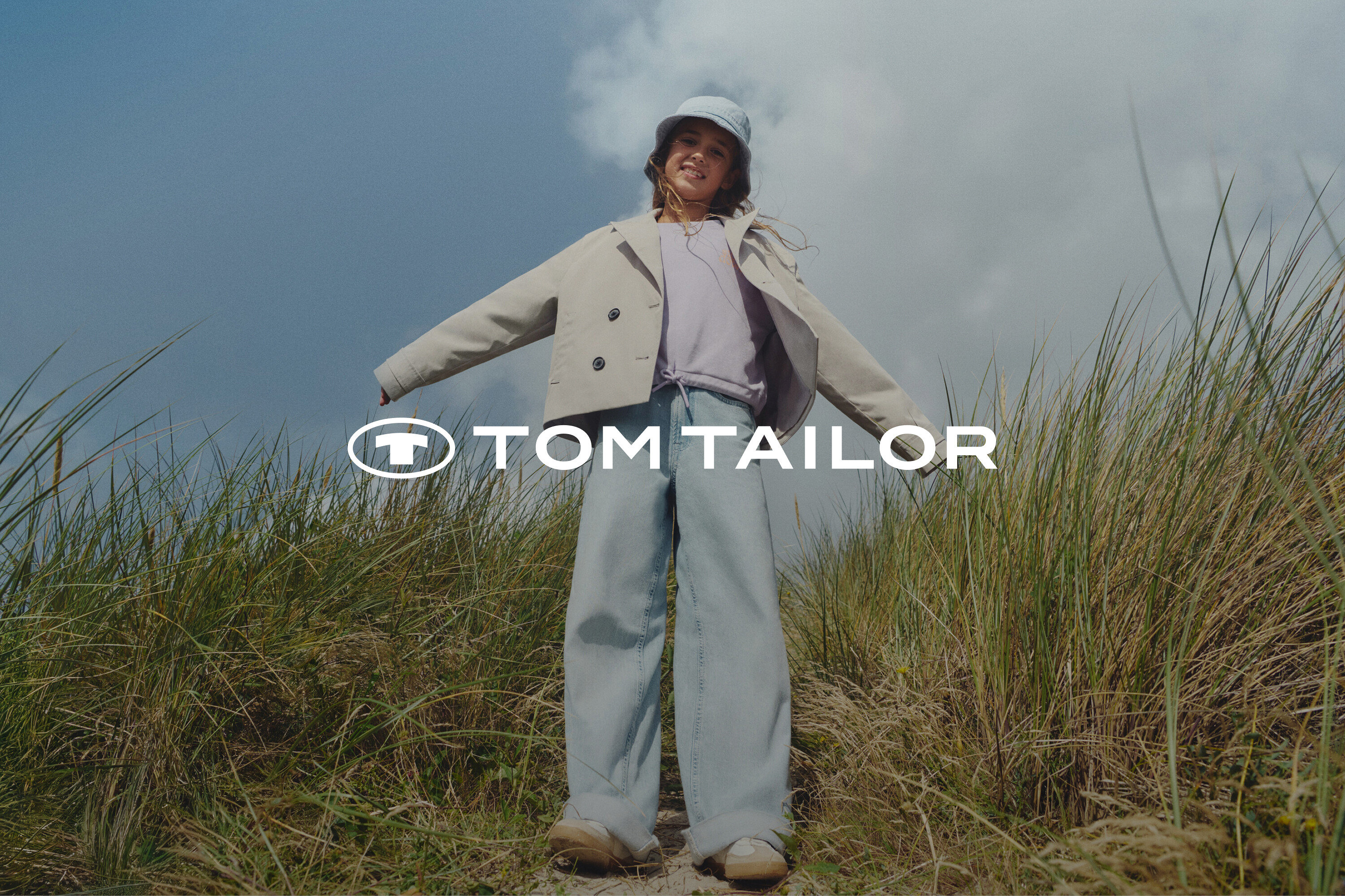 Ein junges Mädchen steht zwischen Gräsern, trägt eine beige Jacke, ein lila Oberteil, weite Jeans und einen hellblauen Hut.