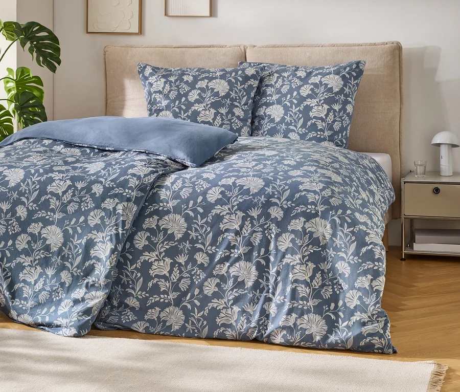 Ein Bett mit blauer Bettwäsche mit weißem Blumenmuster und einem hellblauen Kissen. Das Bett hat ein beiges Kopfteil. Links steht eine Monstera-Pflanze, rechts ein Nachttisch mit Lampe und Glas. Ein beiger Teppich liegt auf dem Holzboden.