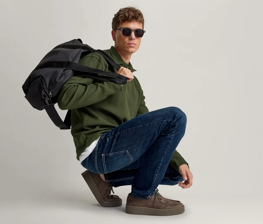 Mann hockt, trägt ein dunkelgrünes Langarm-Poloshirt, blaue Jeans, braune Wildlederschuhe und eine Sonnenbrille, mit einer schwarzen Sporttasche über der linken Schulter.